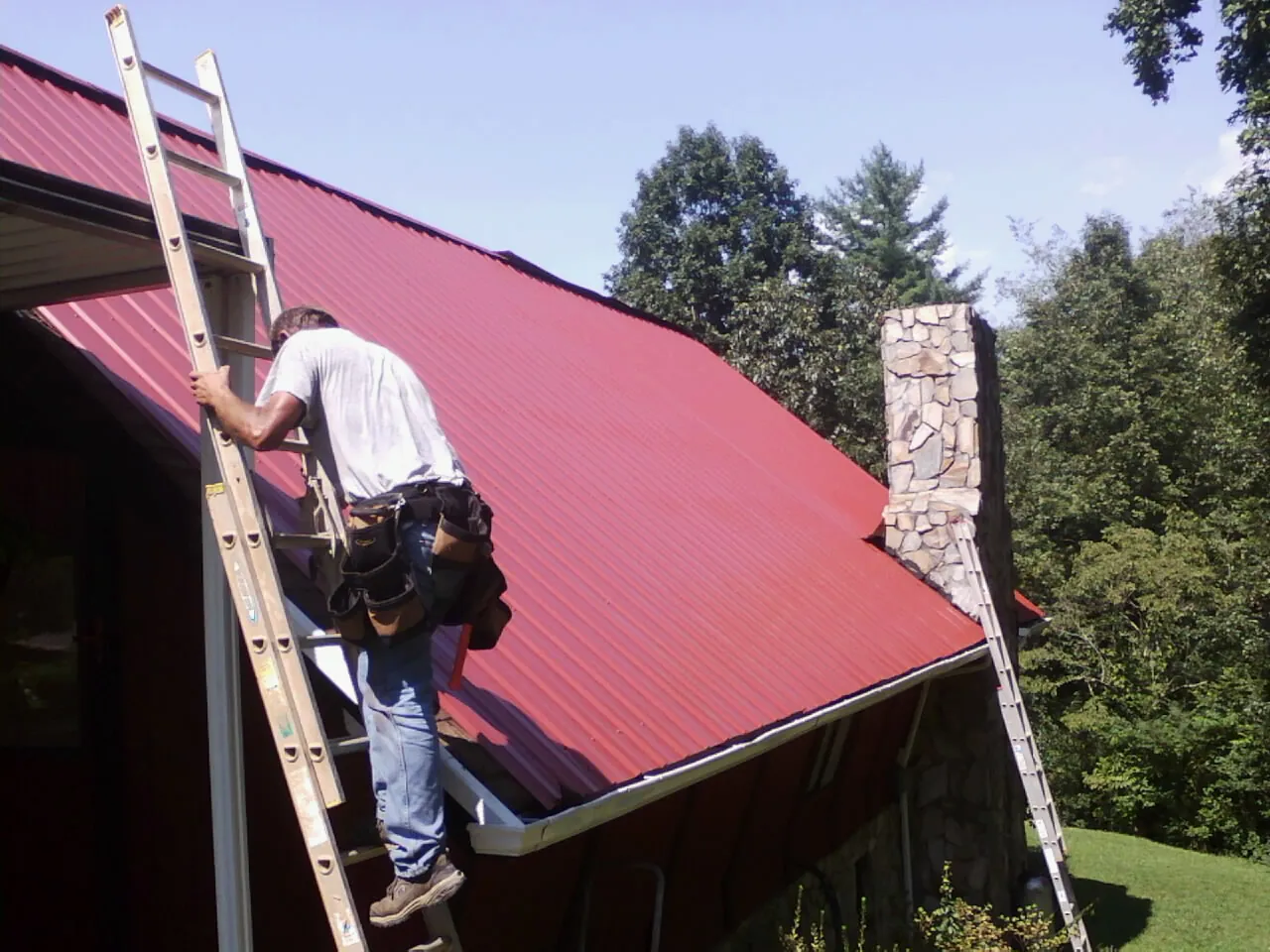 Roofer on a ladder beside a red metal roof and stone chimney — Soffit & Fascia Repair by Roofing Brothers in Crump